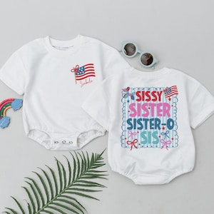 May include: Two white baby bodysuits with patriotic designs. One features an American flag and the name "Isabela." The other has the words "SISSY, SISTER, SISTER-O, SIS" in colorful text, with stars, bows, and flags. A pair of sunglasses and a rainbow are also present.