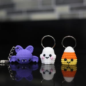 May include: Three crocheted Halloween-themed keychains: a purple bat, a white ghost, and an orange, white, and yellow candy corn. Each keychain has a silver ring.