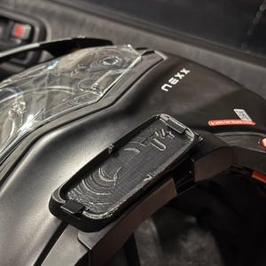 May include: Black motorcycle helmet with a clear visor and a rectangular black accessory attached. The helmet has the word "nexx" printed on the side. A small red sticker is visible.