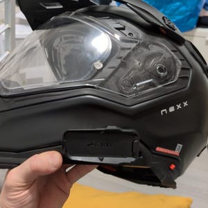Può includere: Un casco da moto nero con una visiera trasparente e un sistema di comunicazione integrato. Il casco ha il nome del marchio "Nexx" stampato sul lato e "Cardo" stampato sul sistema di comunicazione.