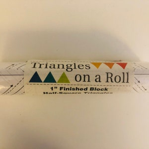 May include: A roll of pre-cut fabric triangles for quilting. The label reads "Triangles on a Roll" with a 1" finished block size and the text "Half-Square Triangles". The label features three triangles in blue, green, and yellow.