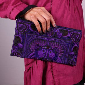May include: A purple embroidered clutch purse with intricate floral and geometric designs. The clutch is made of a dark fabric with a velvet-like texture.