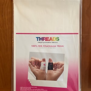 May include: A package of 6 sheets of 13 x 19 inch inkjet printable 100% silk charmeuse fabric. The fabric is white and is packaged in a clear plastic bag. The package has a pink and white label with the text "THREADS inkjet printable fabrics 100% Silk Charmeuse 19mm 6 sheets of 13 x 19" inkjet fabric" and a website address. The image shows a close-up of the fabric with a pair of hands holding two fingers together, with the fingers decorated to look like a bride and groom.