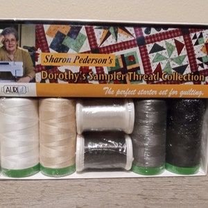 May include: A set of six spools of Aurifil thread in white, beige, gray, and black. The spools are packaged in a clear plastic box with a label that reads "Sharon Pederson's Dorothy's Sampler Thread Collection. The perfect starter set for quilting."