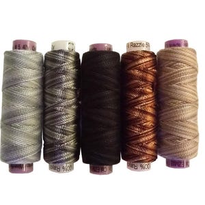 May include: Five spools of thread in various colors, including gray, black, and brown. The spools are labeled with text and appear to be made of rayon. The thread is tightly wound on the spools, and the colors vary from light to dark.