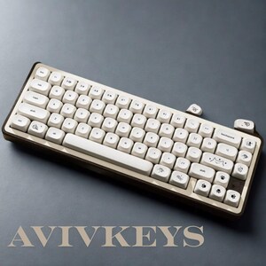 May include: A cream-colored mechanical keyboard with a brown frame. The keyboard has white keycaps with various icons and text. The brand name "AVIVKEYS" is displayed at the bottom. The keyboard is on a gray surface.