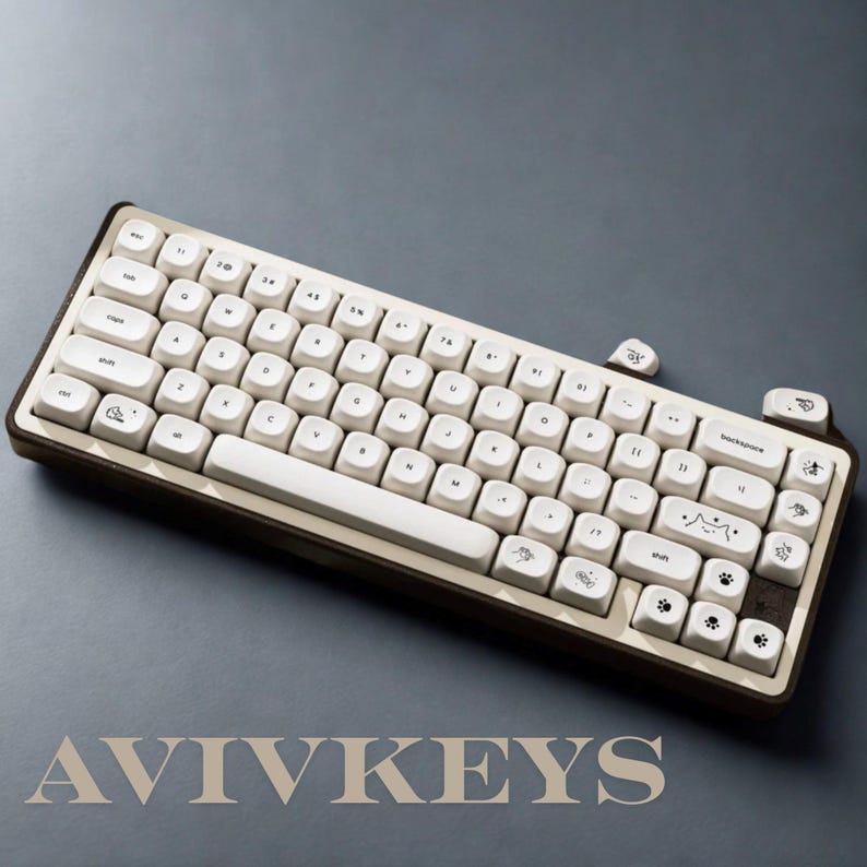 May include: A cream-colored mechanical keyboard with a brown frame. The keyboard has white keycaps with various icons and text. The brand name "AVIVKEYS" is displayed at the bottom. The keyboard is on a gray surface.