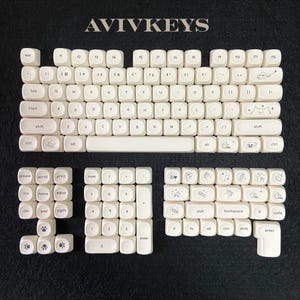 May include: A set of off-white keyboard keycaps with black lettering and whimsical illustrations. The keycaps are arranged on a dark surface, with the brand name "AVIVKEYS" at the top. The set includes various sizes and shapes for a complete keyboard layout.
