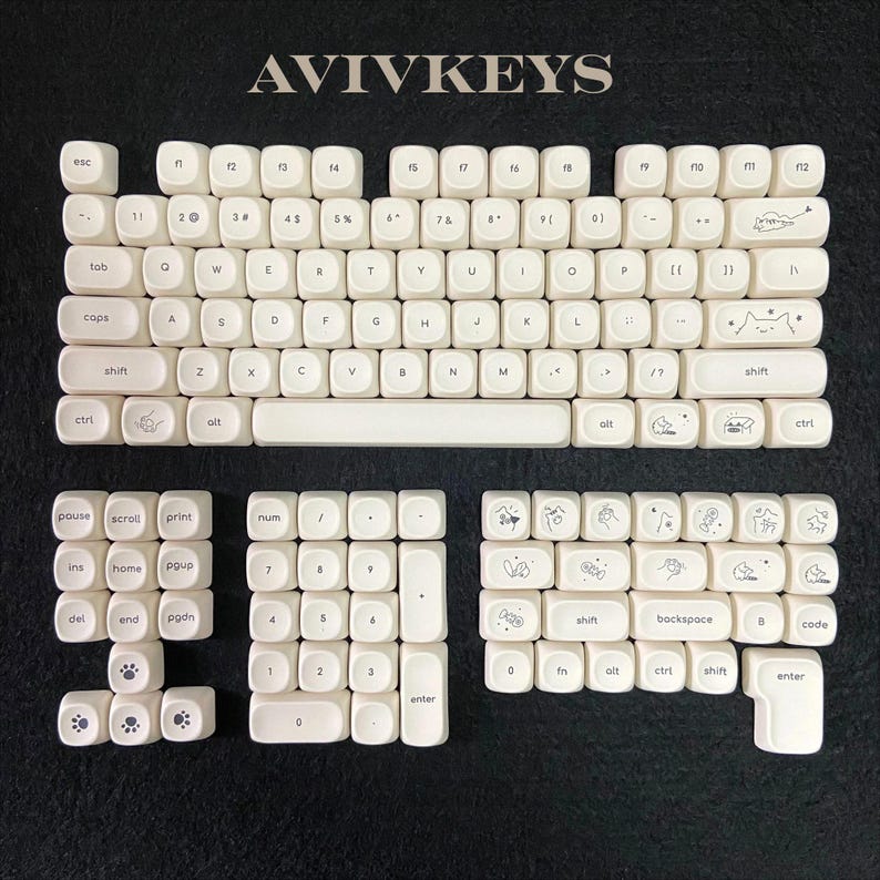 May include: A set of off-white keyboard keycaps with black lettering and whimsical illustrations. The keycaps are arranged on a dark surface, with the brand name "AVIVKEYS" at the top. The set includes various sizes and shapes for a complete keyboard layout.