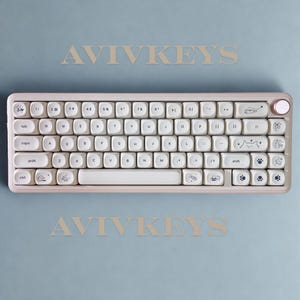May include: A light pink mechanical keyboard with white keycaps. The keyboard has a compact layout with a round volume knob and cat-themed illustrations on some keys. The text "AVIVKEYS" is above and below the keyboard on a light blue background.