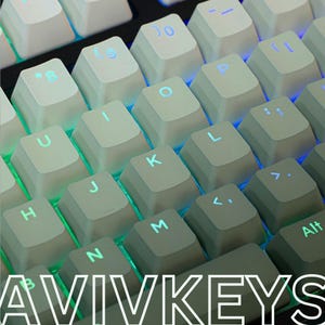 Op de afbeelding: Close-up van een computertoetsenbord met witte keycaps en blauwe en groene achtergrondverlichting. De toetsen zijn gemarkeerd met letters en symbolen. De tekst "AVIVKEYS" staat onderaan.