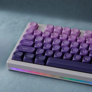 May include: A white mechanical keyboard with purple and lavender keycaps. The keyboard has a gradient color scheme, transitioning from dark purple to light lavender. The keys are arranged in a standard QWERTY layout, with a rainbow LED strip along the bottom edge.