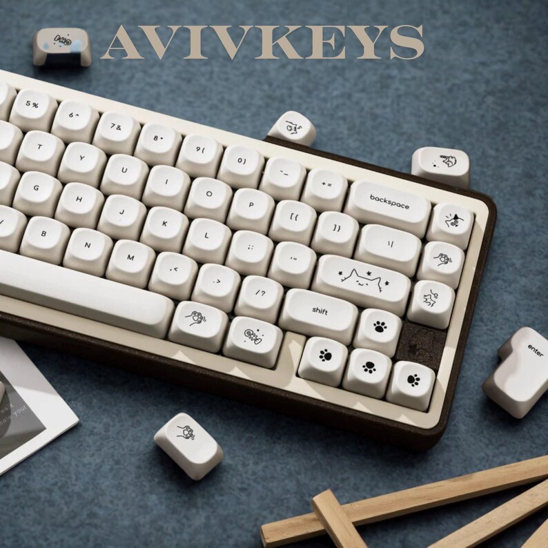 May include: A white and brown mechanical keyboard with custom keycaps. The keycaps feature various illustrations, including paw prints and a cat face. The keyboard has the text "AVIVKEYS" at the top.