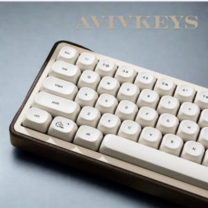 May include: A compact mechanical keyboard with off-white keycaps and a brown frame. The keys are arranged in a standard layout, with the brand name "AVIVKEYS" printed at the top. The keyboard has a minimalist design.