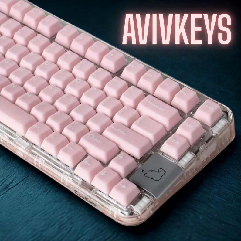 May include: A pink mechanical keyboard with clear casing. The keyboard has pink keycaps and the text "AVIVKEYS" in the upper right corner. The keyboard is on a dark blue surface.