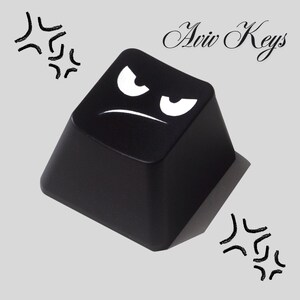 May include: A black keycap with a white angry face design. The keycap has the text "Aviv Keys" in cursive. The background is a light gray color with black graphic elements.