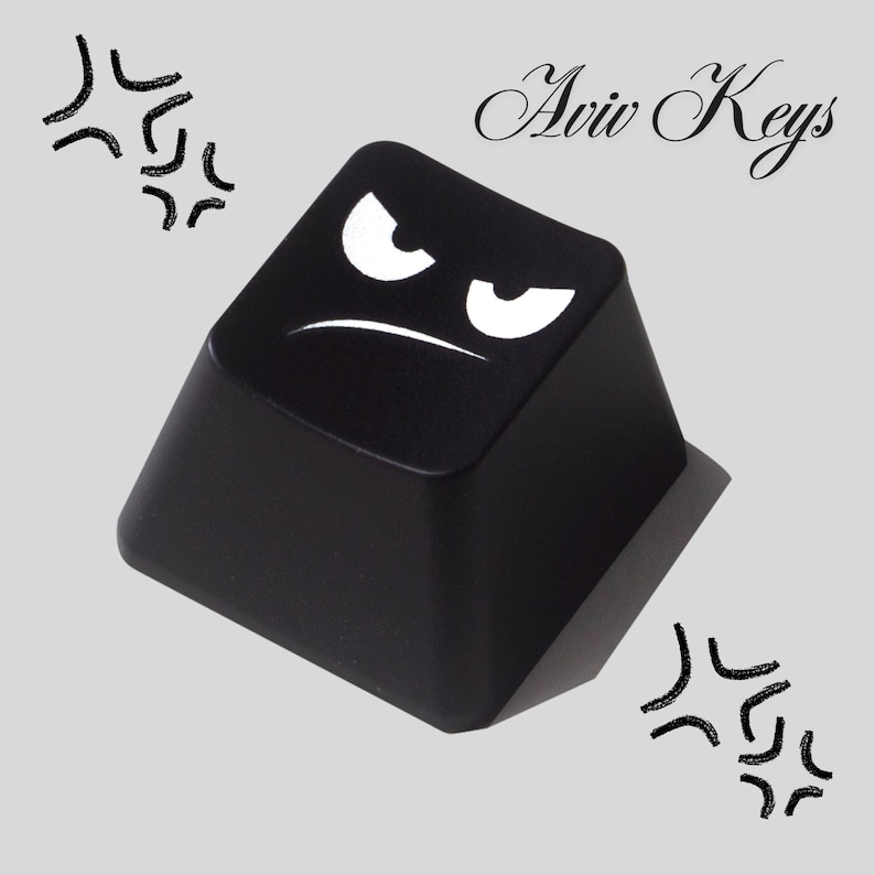 May include: A black keycap with a white angry face design. The keycap has the text "Aviv Keys" in cursive. The background is a light gray color with black graphic elements.