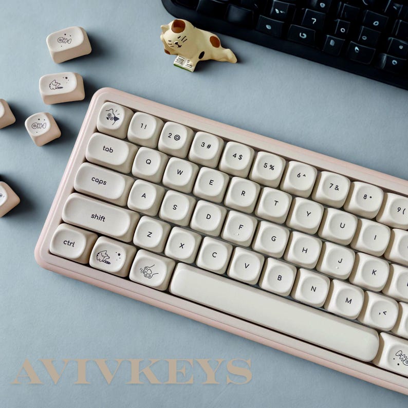 May include: A cream-colored mechanical keyboard with a pink frame and custom keycaps featuring cartoon illustrations. A small cat figurine and extra keycaps are placed nearby. The keyboard has a standard QWERTY layout.