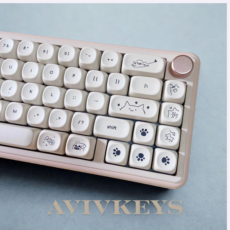 May include: A light pink and white mechanical keyboard with custom keycaps. The keycaps feature cat-themed illustrations and paw prints. The keyboard has a rose gold-colored knob and the text "AVIVKEYS" at the bottom.