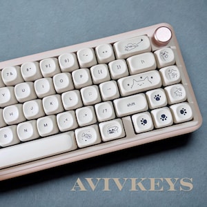 May include: A cream-colored mechanical keyboard with custom keycaps featuring cat and paw print designs. The keyboard has a rose gold accent and the text "AVIVKEYS" is visible at the bottom.
