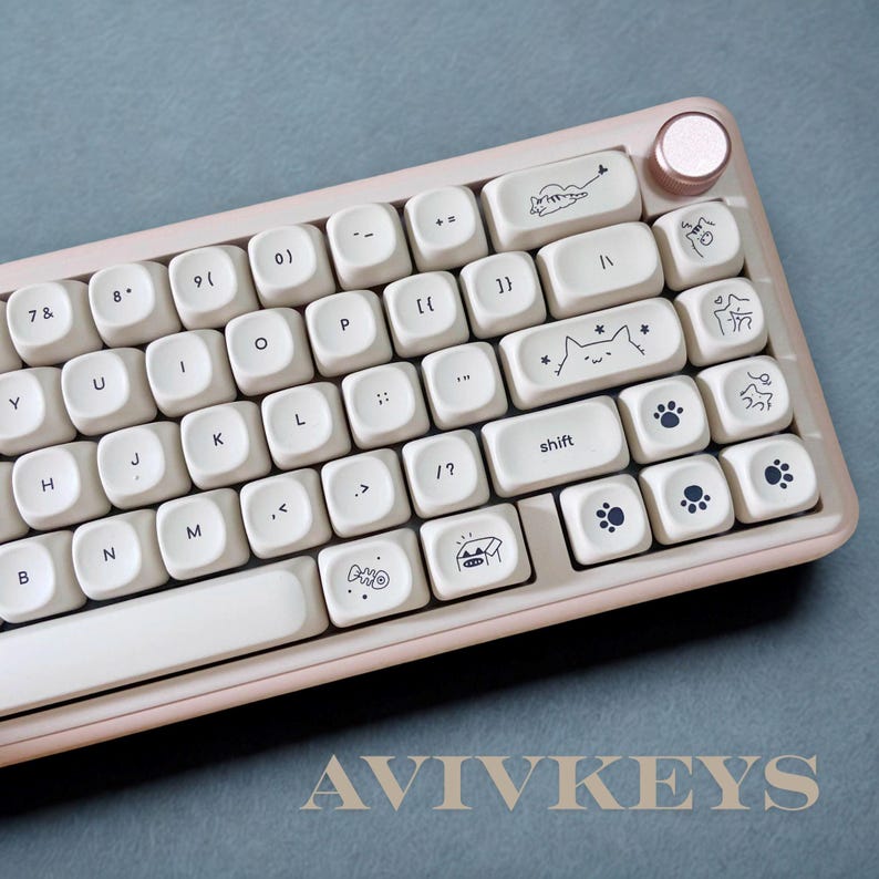 May include: A cream-colored mechanical keyboard with custom keycaps featuring cat and paw print designs. The keyboard has a rose gold accent and the text "AVIVKEYS" is visible at the bottom.