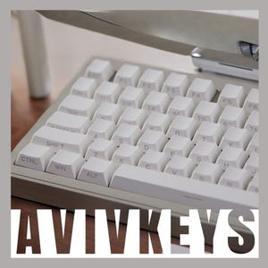 May include: A white computer keyboard with a white monitor in the background. The keyboard has a white frame and white keys with black lettering. The keys are labeled with letters, numbers, and symbols. The keyboard has a row of function keys at the top, a row of number keys, and a row of letter keys. The keyboard also has a row of control keys, including Ctrl, Win, and Alt. The keyboard is on a wooden surface. The text "AVIVKEYS" is printed on the surface below the keyboard.
