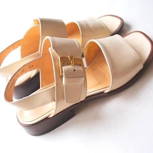 May include: Cream-colored leather sandals with a square toe and a strap that buckles with a gold-colored metal clasp. The sandals have a low heel and a dark brown sole. The sandals are shown on a white surface.