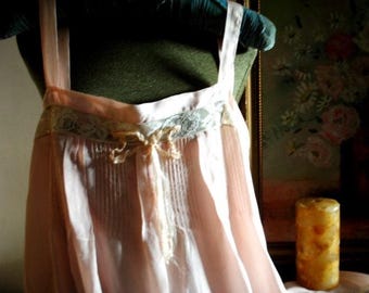 May include: A pale pink sleeveless garment featuring a horizontal band of light green lace across the chest, vertical pleats below the neckline, and a small fabric bow centered at the front. The piece has wide shoulder straps and a loose silhouette.