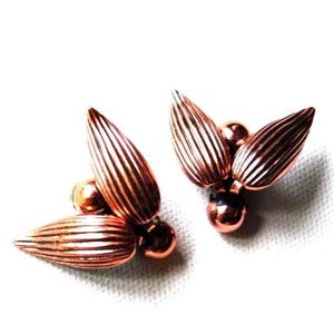 May include: A pair of copper-colored earrings. Each earring features three stylized, ribbed petal shapes radiating from a central sphere. The earrings have a polished, metallic finish and are set against a white background.