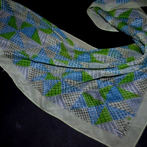 May include: A white silk scarf with a geometric pattern of green, blue, and black squares and triangles. The scarf has a fringed edge and the text "Vera" is printed in the lower right corner.