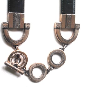 Peut inclure: Une ceinture en cuir noir avec des d&eacute;tails en m&eacute;tal dor&eacute;. La ceinture est dot&eacute;e d'une boucle d&eacute;corative avec un motif orn&eacute; et d'une fermeture &agrave; cha&icirc;ne.