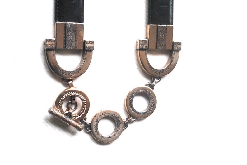 Peut inclure: Une ceinture en cuir noir avec des d&eacute;tails en m&eacute;tal dor&eacute;. La ceinture est dot&eacute;e d'une boucle d&eacute;corative avec un motif orn&eacute; et d'une fermeture &agrave; cha&icirc;ne.