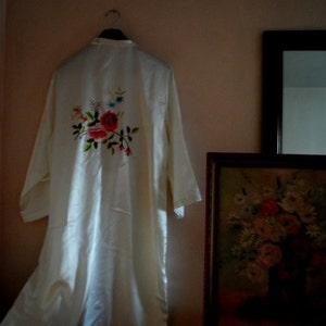 Könnte beinhalten: Eine weiße Satin-Robe mit gestickten Blumenmotiven. Die Stickerei zeigt eine rosa Rose, grüne Blätter und weiße Blüten.