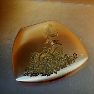 May include: A gold-colored compact with a floral design and a raised, stylized bird motif.