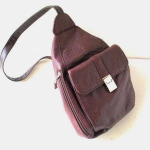 May include: A burgundy leather sling bag with a long, adjustable strap. The bag has a front flap pocket with a silver-coloured metal clasp and a zipped compartment. The bag is designed to be worn across the body.