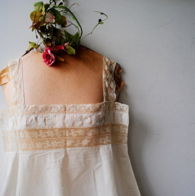 May include: A white cotton slip with delicate lace trim. The slip is on a dress form with a floral decoration.