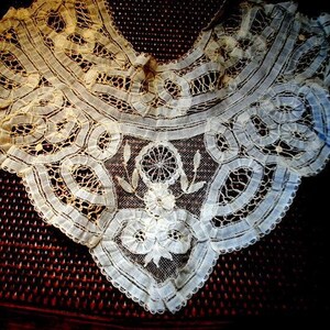 May include: An antique, cream-coloured lace collar with detailed floral and geometric patterns. The delicate lace features a scalloped edge and a central floral motif. The collar is displayed on a dark, textured surface, highlighting its fine craftsmanship.