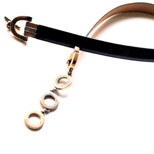 Peut inclure: Une ceinture en cuir noir avec une boucle dor&eacute;e et une cha&icirc;ne avec trois anneaux dor&eacute;s qui pendent.