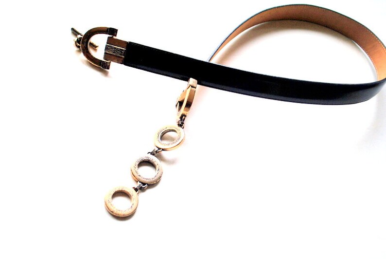 Peut inclure: Une ceinture en cuir noir avec une boucle dor&eacute;e et une cha&icirc;ne avec trois anneaux dor&eacute;s qui pendent.