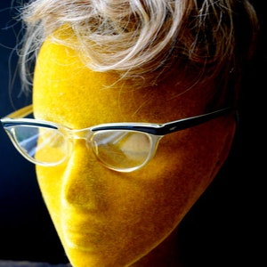 Könnte beinhalten: Ein gelber Mannequin-Kopf mit blonden Haaren, der eine klare Cat-Eye-Brille mit schwarzen Rahmen trägt.