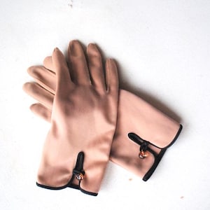 May include: A pair of light brown gloves with black trim and decorative buttons. The gloves are made of a soft, smooth fabric.
