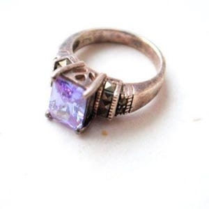 May include: A silver ring with a square, light purple gemstone. The ring has a decorative setting with smaller, dark stones on the sides. The band is a simple, rounded design.