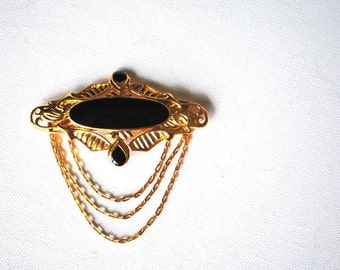 Art deco vintage 80s, gold tone metal brooch with a black lucite , oval shape centerpiece and three small , dangle chains drapery . Trifary.