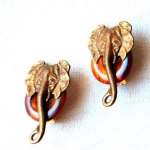 Può includere: Un paio di orecchini vintage a forma di testa di elefante dorata. Gli elefanti hanno dettagli e sono montati su una base circolare marrone e ambrata. Orecchini a clip.