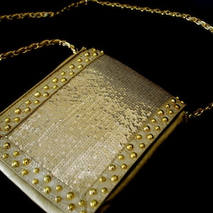 May include: A gold sequined clutch bag with a gold chain strap and gold studs around the edges.