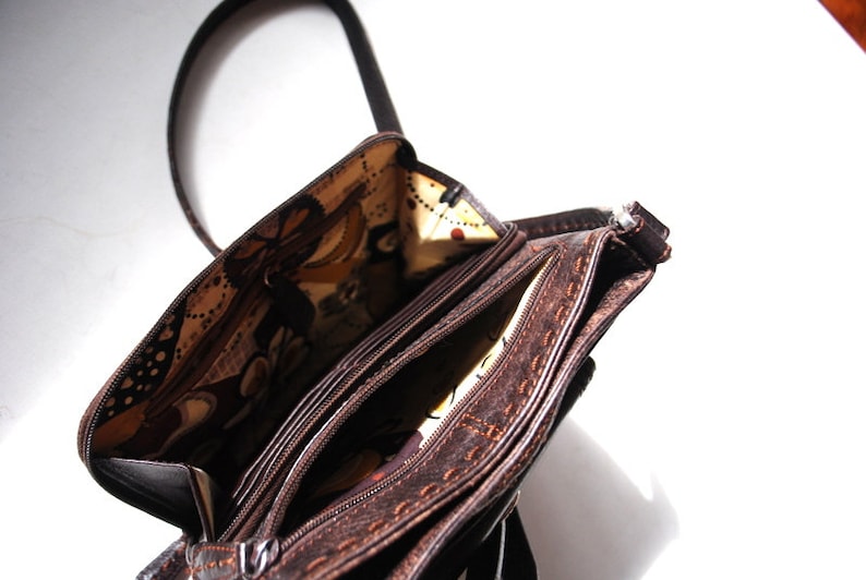 May include: A brown leather purse with a zipper closure and a patterned fabric lining. The purse has a long strap and multiple compartments.