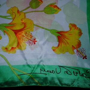 Puede incluir: Pañuelo de seda con un diseño floral con flores de hibisco amarillas y naranjas y hojas verdes. El pañuelo tiene un fondo blanco con un borde verde y una firma en negro.