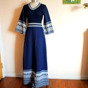 May include: A navy blue maxi dress with bell sleeves and a geometric pattern on the cuffs and hem. The dress features a white and navy trim around the neckline and waist. The dress is displayed on a mannequin.