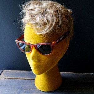 May include: A pair of vintage-style sunglasses with a tortoiseshell frame and dark lenses rests on a yellow mannequin head with blonde hair. The sunglasses have a classic, retro design, perfect for adding a touch of style.