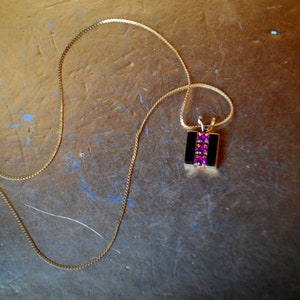 May include: A gold chain necklace with a pendant featuring a black rectangular stone with a row of pink gemstones.